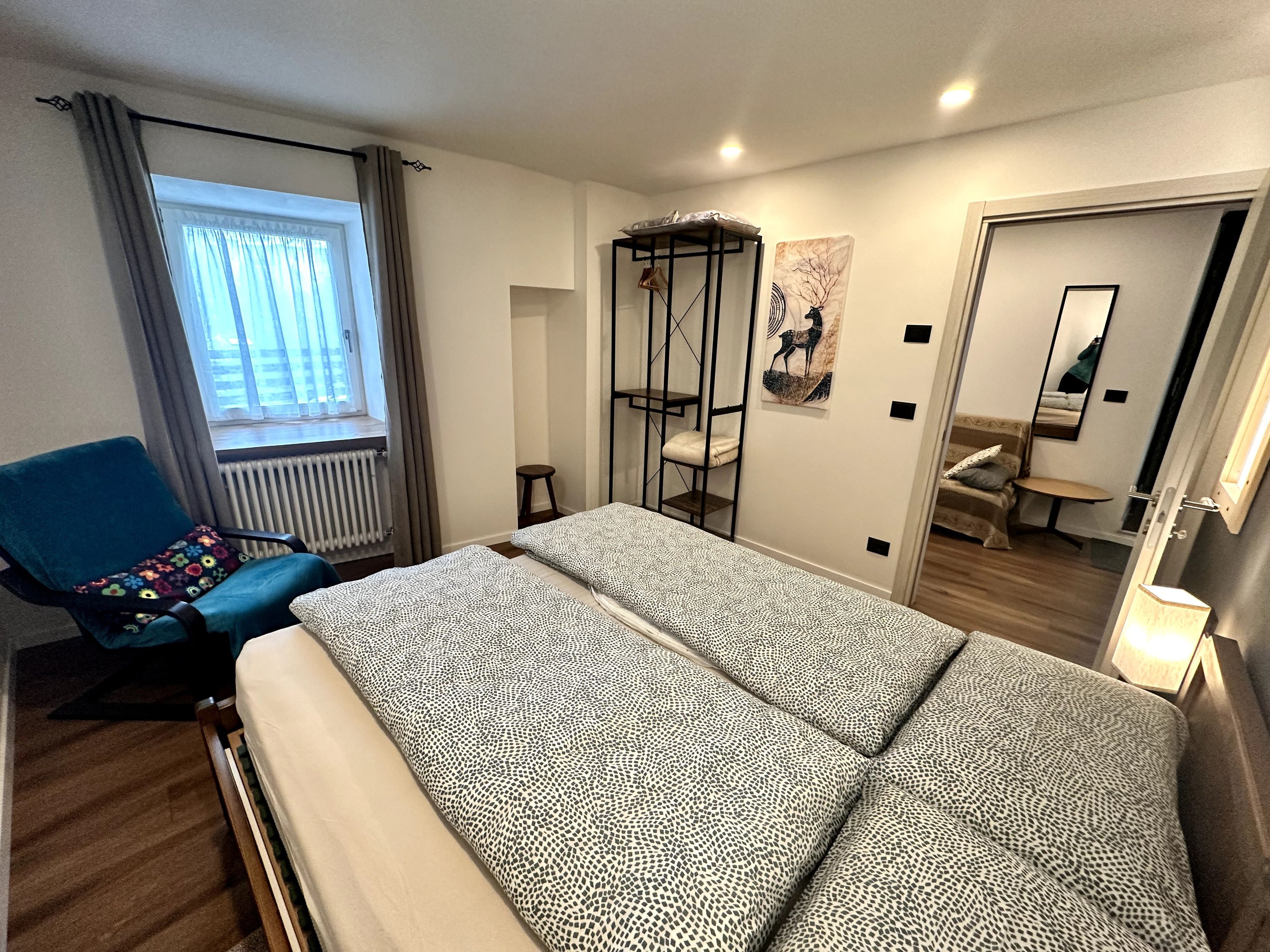 Ein Schlafzimmer mit Bett, Sessel, Fenster mit Vorhängen, Heizkörper, Hocker, Wandregal, Spiegel und Tür.