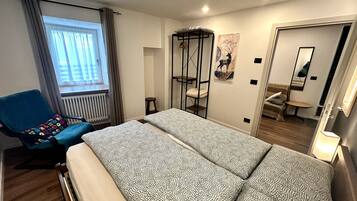 Ein Schlafzimmer mit Bett, Sessel, Fenster mit Vorhängen, Heizkörper, Hocker, Wandregal, Spiegel und Tür.