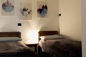 A room with two single beds, a nightstand with a lamp, and three wall art pieces depicting nature scenes.