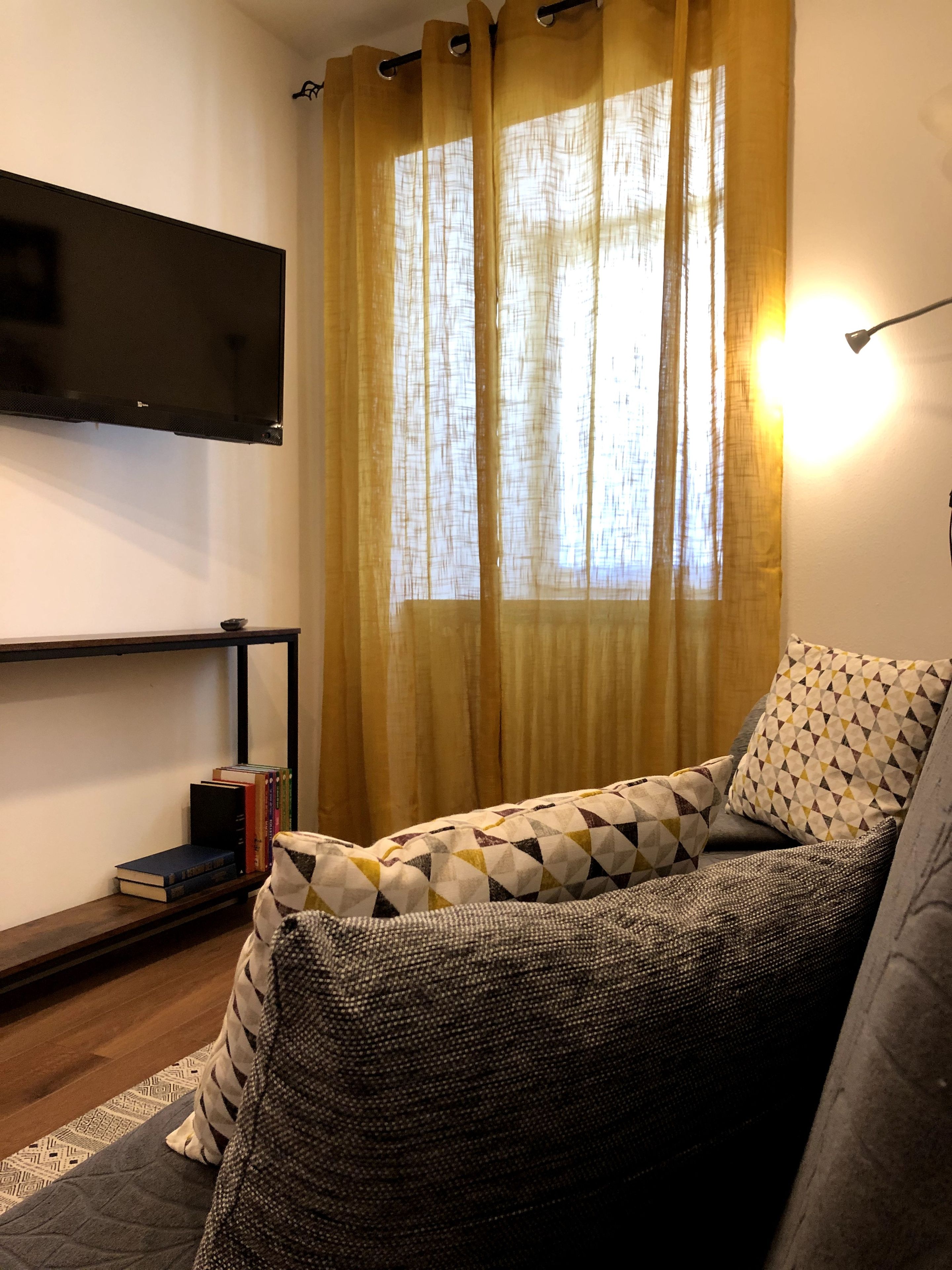 Ein Wohnzimmer mit einer Couch, einem flachbildfernseher an der Wand, einem Bücherregal mit Büchern und einem Fenster mit Vorhängen.