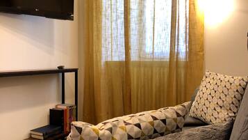Ein Wohnzimmer mit einer Couch, einem flachbildfernseher an der Wand, einem Bücherregal mit Büchern und einem Fenster mit Vorhängen.