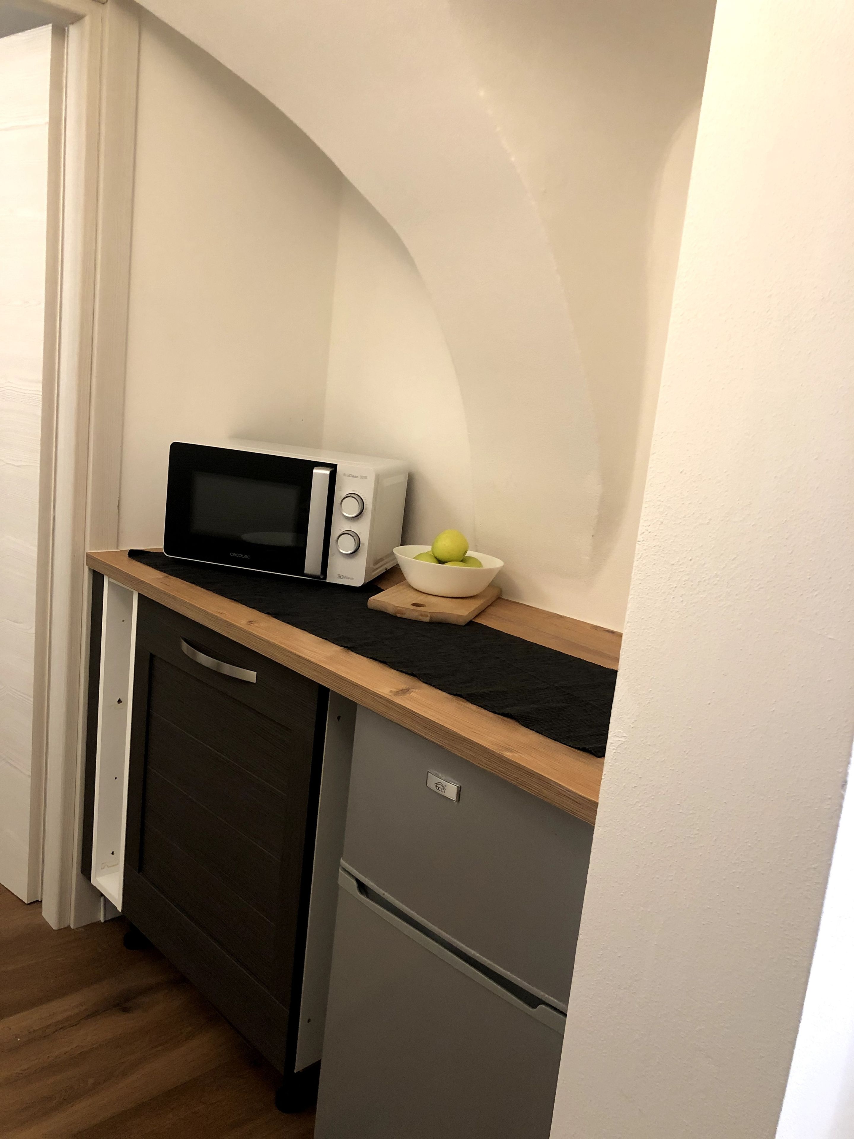 Eine kompakte Küchenzeile mit einer Mikrowelle, einem kleinen Kühlschrank und einer Schüssel Äpfel auf der Arbeitsfläche.