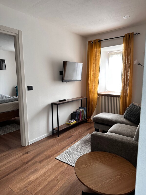 A modern living room with a sofa, a small table, a TV mounted on the wall, and a window with curtains.