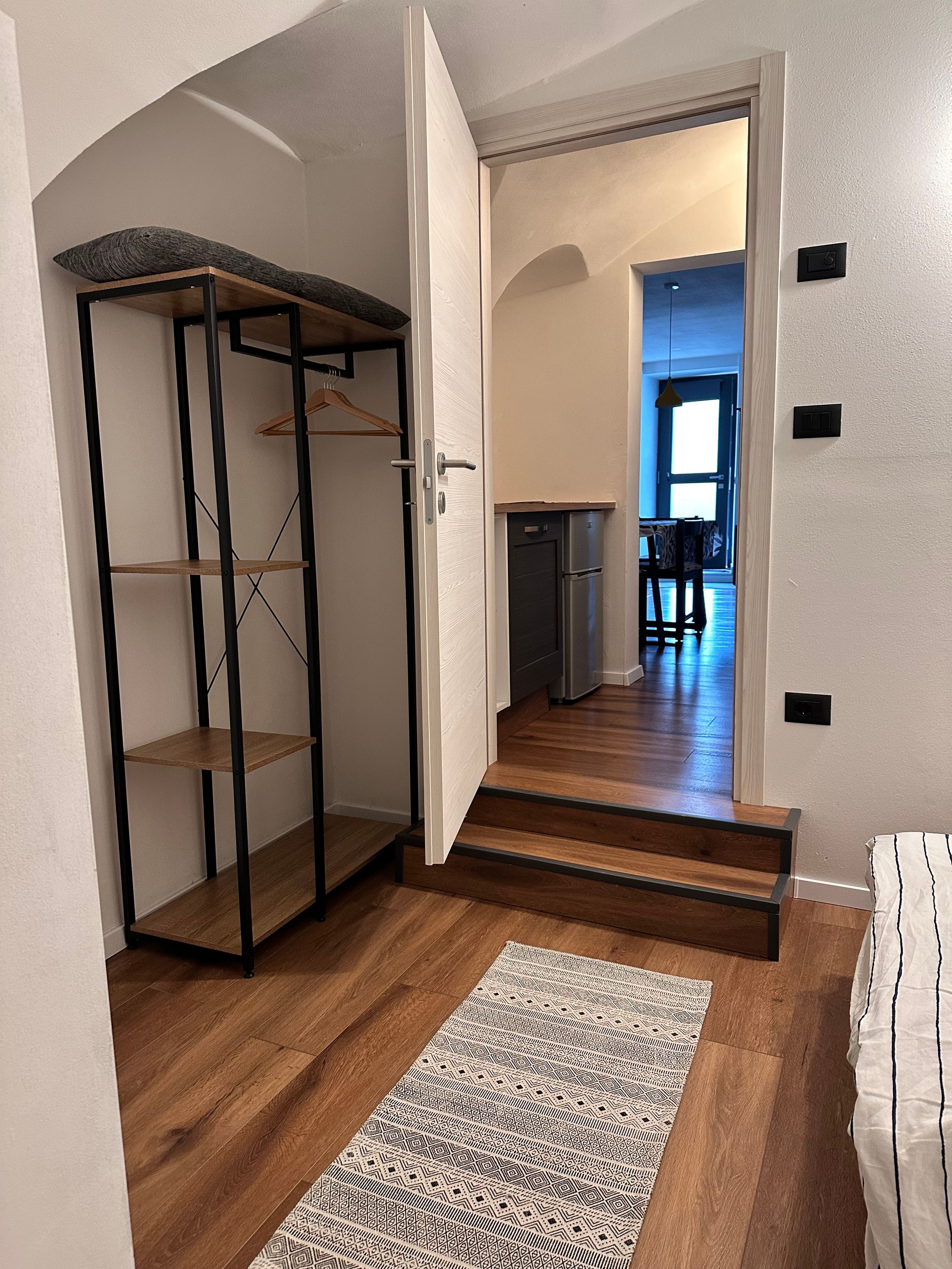 Ein Flur mit Holzboden, einem gemusterten Teppich, einem Metallregal und einer Tür zu einem Küchenbereich.
