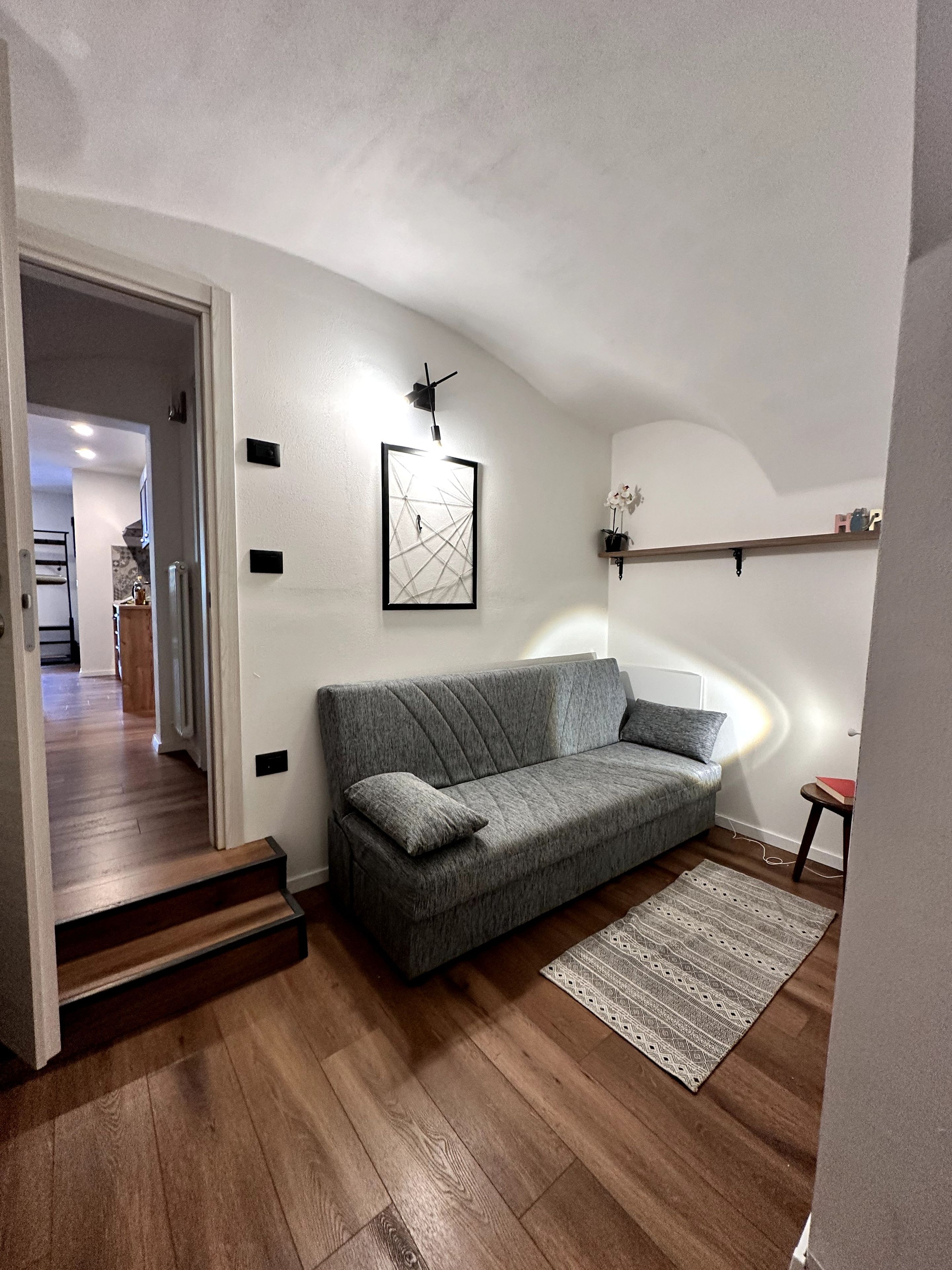 Una sala moderna con un sofá gris, una obra de arte abstracta enmarcada, una mesita con un libro y una escalera que lleva a otra habitación.