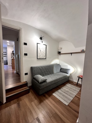 Ein modernes Wohnzimmer mit einem grauen Sofa, einem gerahmten abstrakten Kunstwerk, einem kleinen Tisch mit einem Buch und einer Treppe, die in einen anderen Raum führt.