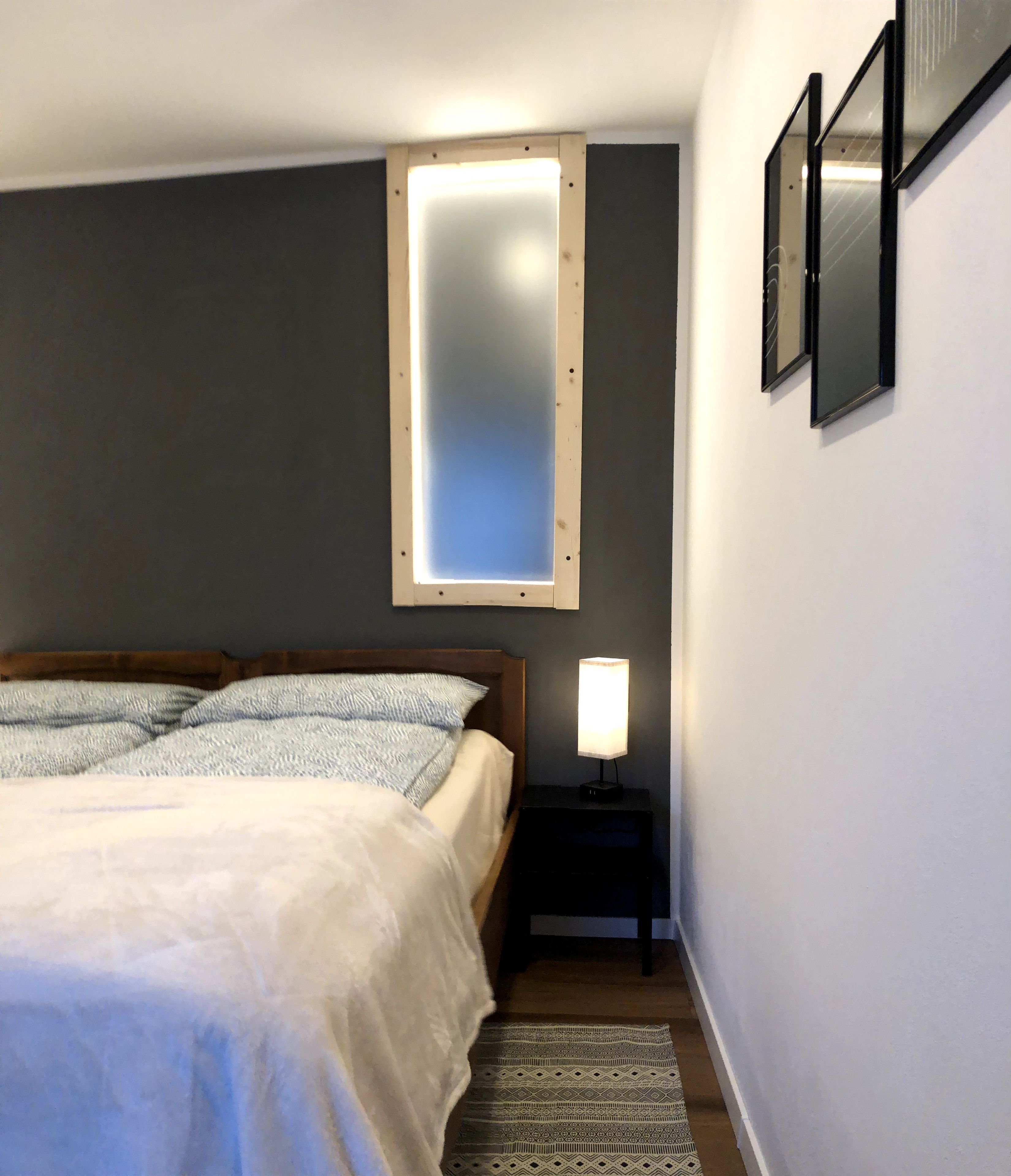 Ein Schlafzimmer mit einem Bett, einem Nachttisch mit Lampe, einem Fenster und einem Wandspiegel.