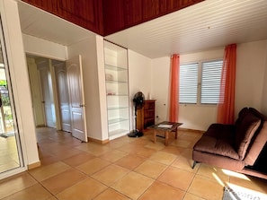 A living room with a brown sofa, a glass shelving unit, a fan, and a window with blinds. - Fare Arava Huahine - 2-Bedroom Accommodation 50 Meters from the Beach (Fare)