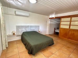 A room with a bed, a nightstand, a lamp, an air conditioner, and a sliding door leading to another room.