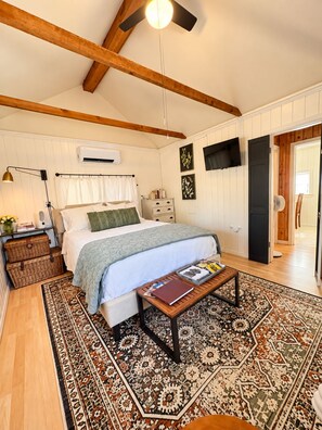 Ein Schlafzimmer mit einem Bett, Nachttischen, einem Schreibtischchen, einem Deckenventilator, einer Klimaanlage, einem Fernseher und einem gemusterten Teppich.