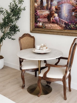 A round table with a white marble top, surrounded by two upholstered chairs with wooden frames and a woven backrest. A small white table with a few items is placed on the table.