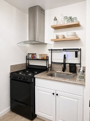 A kitchen with a black stove, white cabinets, a stainless steel range hood, a granite countertop, a black faucet, and a wooden shelf above the sink.