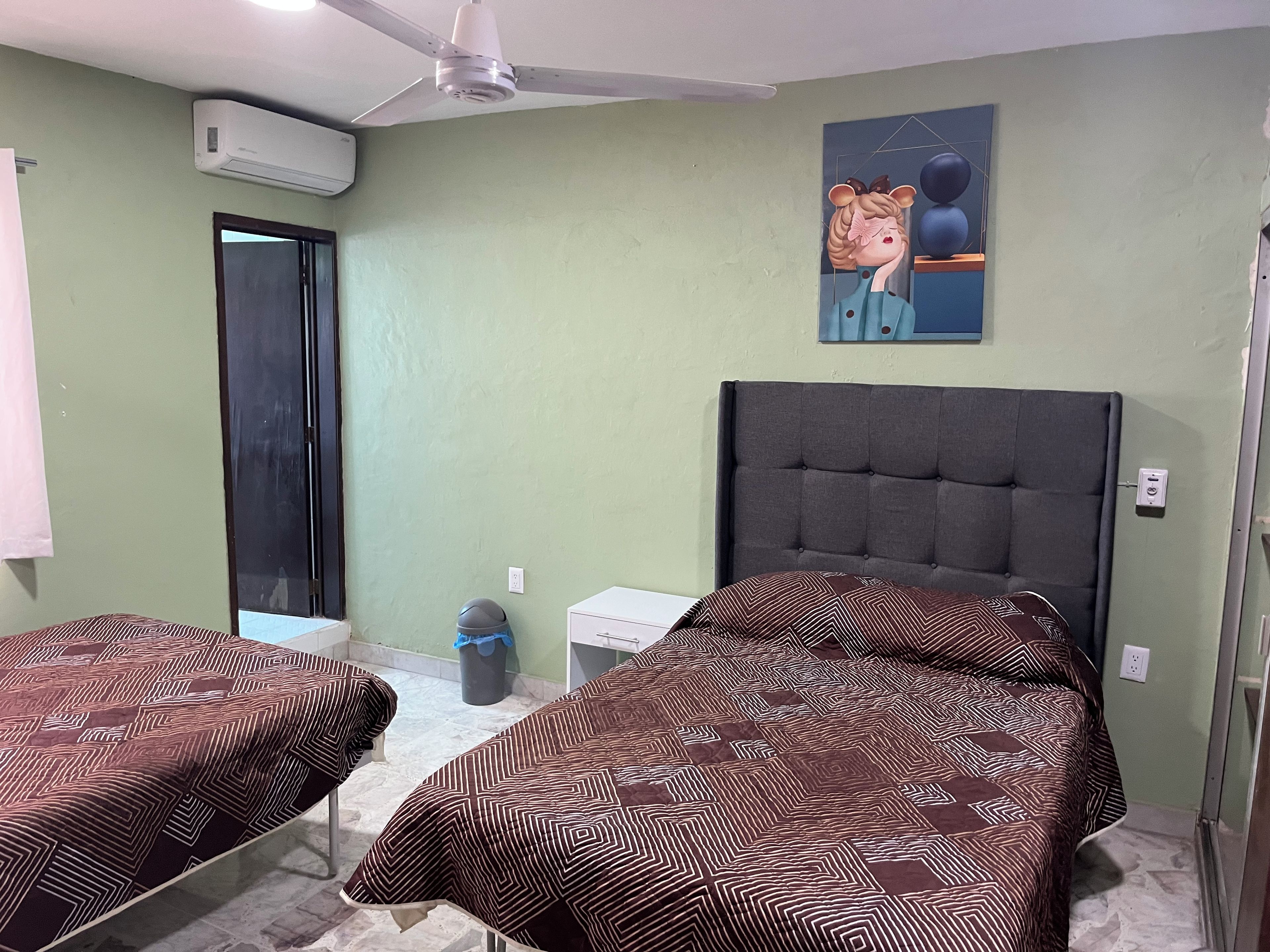 Une chambre avec deux lits, un ventilateur de plafond, un appareil de climatisation, une poubelle et un tableau au mur.