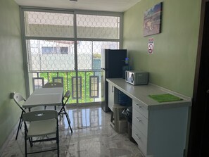 A room with a dining table and chairs, a microwave on a counter, a water dispenser, and a view through a window with a metal fence.