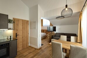 A modern kitchen with a built-in oven, a dining area with a wooden table and chairs, a living room with a sofa and a TV, and a staircase leading to an upper level. - Penthouse Suite With Panoramic Views (Gaschurn)