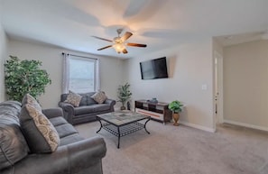 A living room with a ceiling fan, a grey sofa, a coffee table, a TV mounted on the wall, and a door to the right.