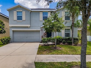 A two-story residential building with a garage, a small tree in the front yard, and a clear sky. - Spacious 5-bedroom house in peaceful Wimauma with AC (Wimauma)