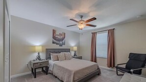 Un dormitorio con cama, dos mesitas de noche, ventilador de techo, ventana con cortinas y una silla.