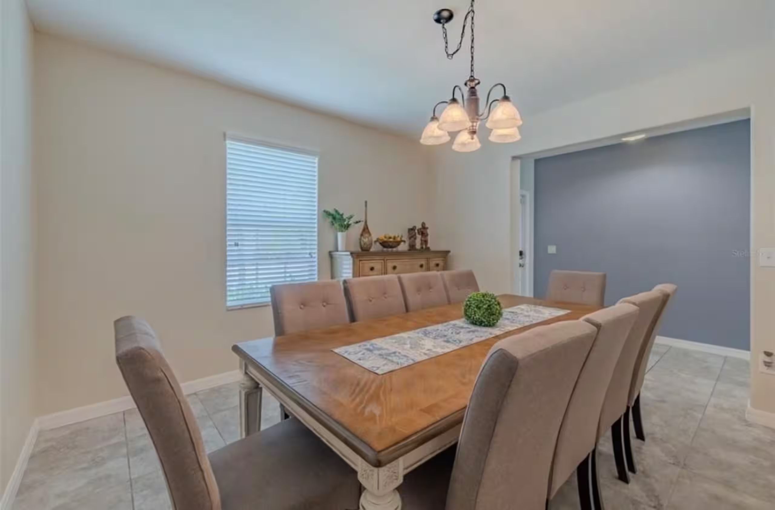 A dining room with a wooden table, six chairs, a chandelier, a window with blinds, and a door.
