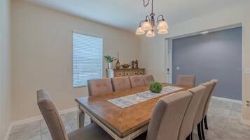 A dining room with a wooden table, six chairs, a chandelier, a window with blinds, and a door.