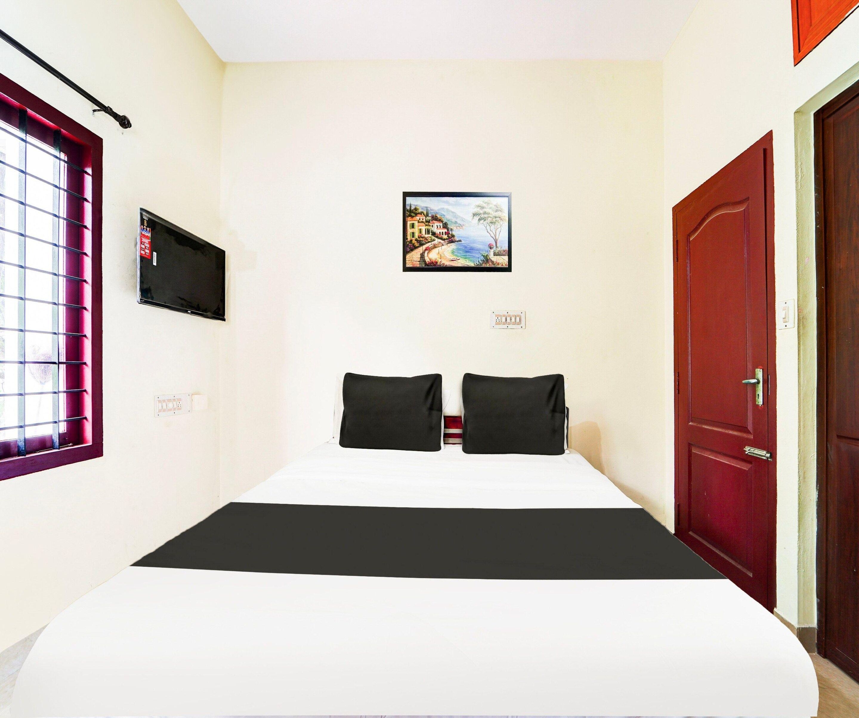 Una habitación de hotel con una cama, un televisor montado en la pared, un cuadro sobre la cama y una puerta roja.