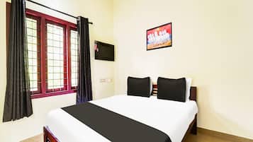 A hotel room with a bed, curtains, a window with bars, a wall-mounted TV, and a painting.