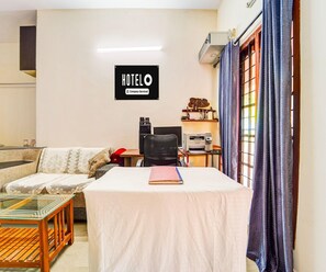 Een hotelkamer met een bank, een tafel met een boek, een bureau met een computer en een printer, en een bordje waarop staat dat het een bedrijfsdienstverlening betreft.