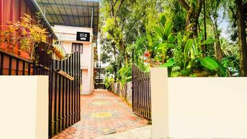 A hotel entrance with a paved pathway leading to a gated entrance, surrounded by lush greenery and trees.