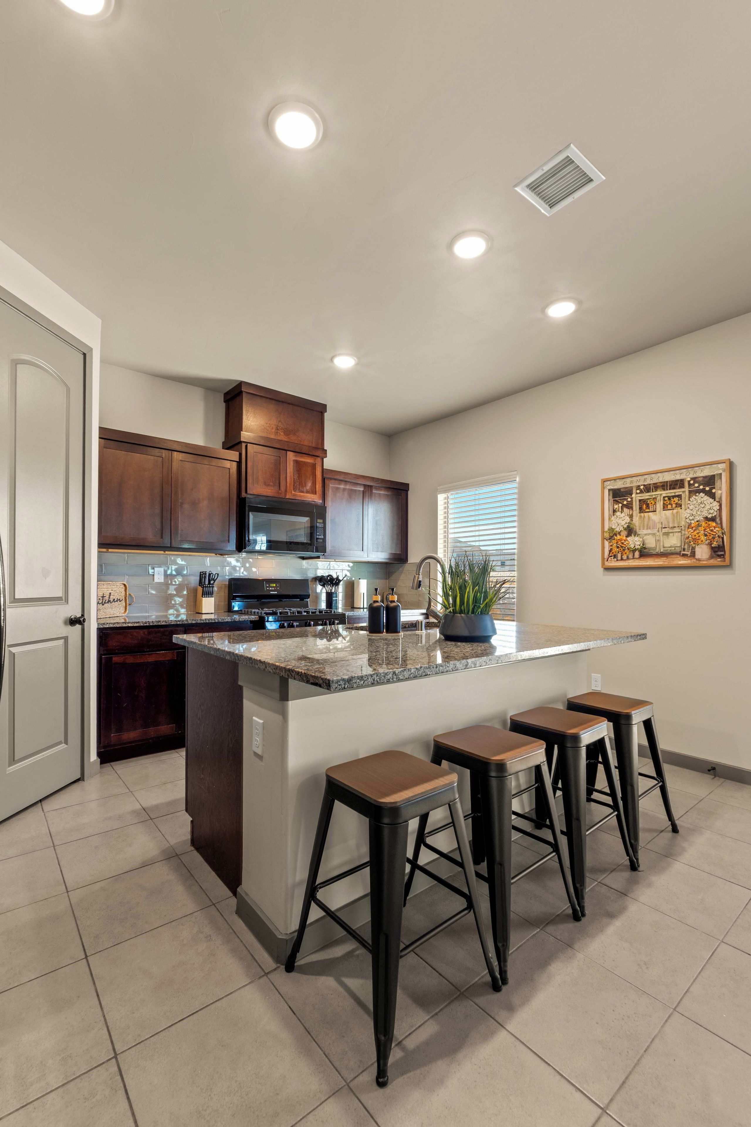 A kitchen with a central island, bar stools, wooden cabinets, a microwave, and a window with blinds.