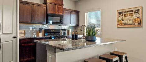 A kitchen with a central island, bar stools, wooden cabinets, a microwave, and a window with blinds.
