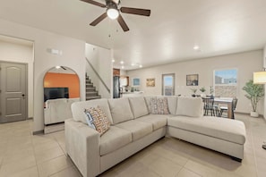A modern living room with a sectional sofa, a ceiling fan, a staircase, and a dining area.