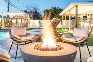 A backyard with a fire pit surrounded by wicker chairs and a covered patio area.