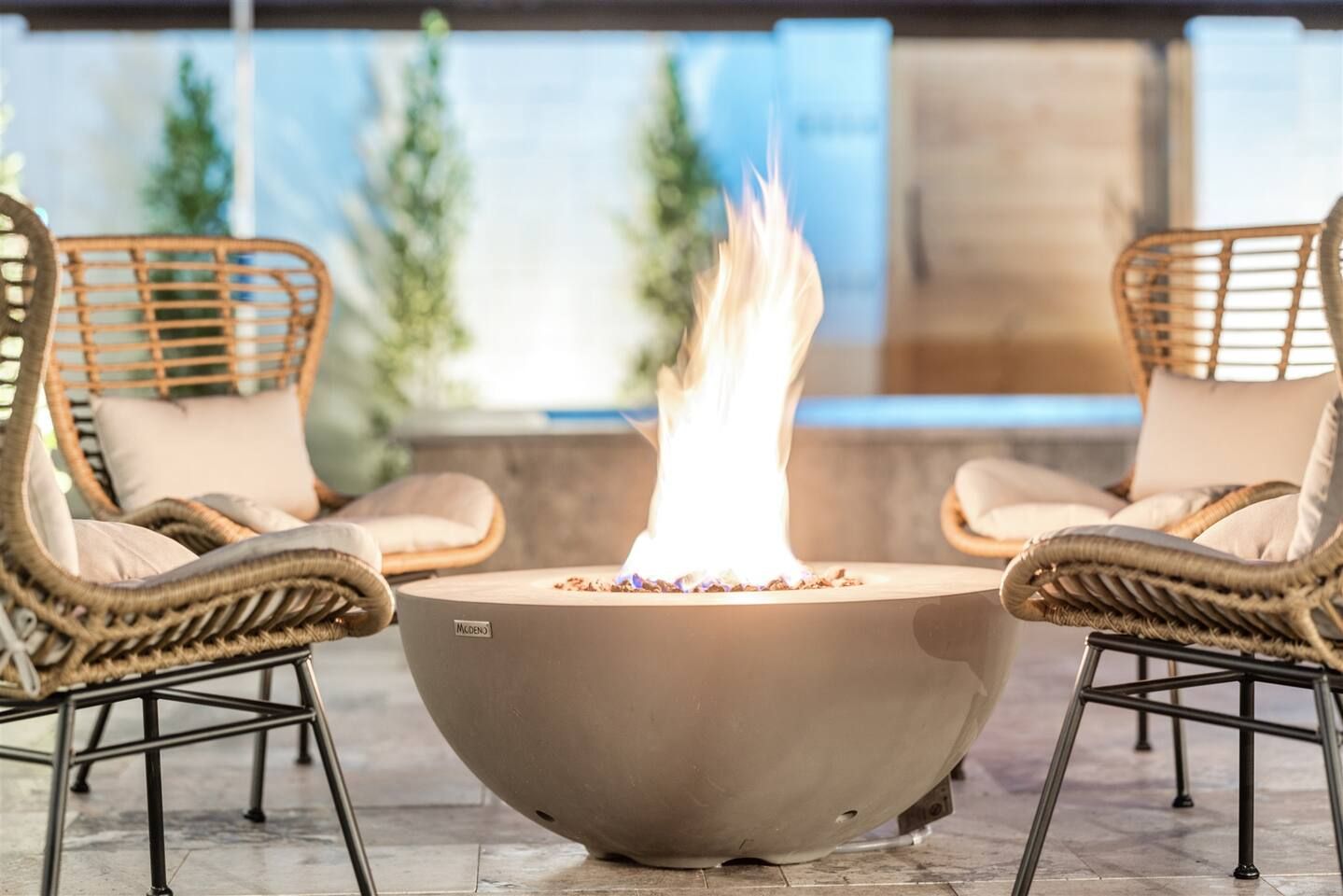 Un foyer extérieur de forme ronde, doté d’une structure métallique et entouré de fauteuils en osier avec des coussins.