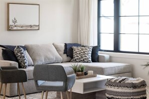 A modern living room with a grey sofa, patterned cushions, a small side table with a plant, and a window with curtains. - Reesor: Scottsdale Home with Private Pool & Spa (Scottsdale)
