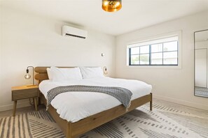 A bedroom with a wooden bed, white bedding, a nightstand, a lamp, an air conditioning unit, a window with blinds, and a mirror. - Reesor: Scottsdale Home with Private Pool & Spa (Scottsdale)