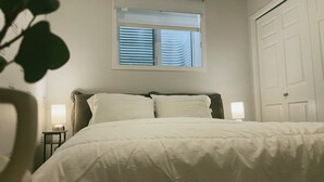 A bed with a white comforter, a window with blinds, a bedside lamp, and a door. - Modern New 2BR beside Park, 14m WEM, 4m Grocery (Edmonton)