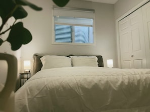 Ein Bett mit weißer Bettdecke, ein Fenster mit Jalousien, eine Nachttischlampe und eine Tür.
