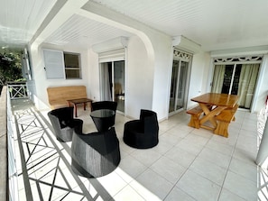 A covered outdoor seating area with a wooden table and chairs, a bench, and a glass-top table.