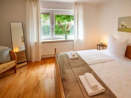 Habitación de hotel con cama, una silla, mesita de noche con lámpara, ventana con cortinas y vistas a zonas verdes.