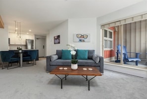 A modern living room with a grey sofa, a wooden coffee table, and a dining area with a black table and blue chairs. - Creekside Gondola, pool, hot tub, BBQ, & free parking! (Whistler)