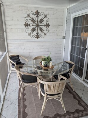 Une table à plateau en verre, quatre chaises en osier, un élément décoratif mural et un panneau d’accueil sur un porche.