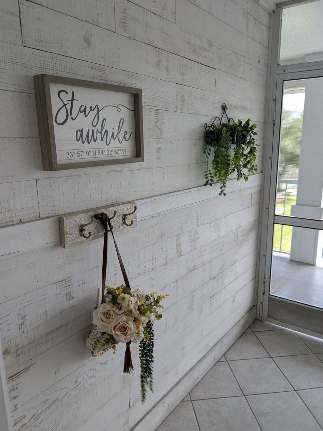 A wall with a framed sign saying 'Stay awhile' and coordinates, a hanging plant, and a floral arrangement.