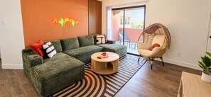 A modern living room with a green sofa, a round coffee table, a wicker chair, and a neon light on the wall. - The Saguaro - King • Arcade • Pool/Spa • Gym • W/D (Tempe)