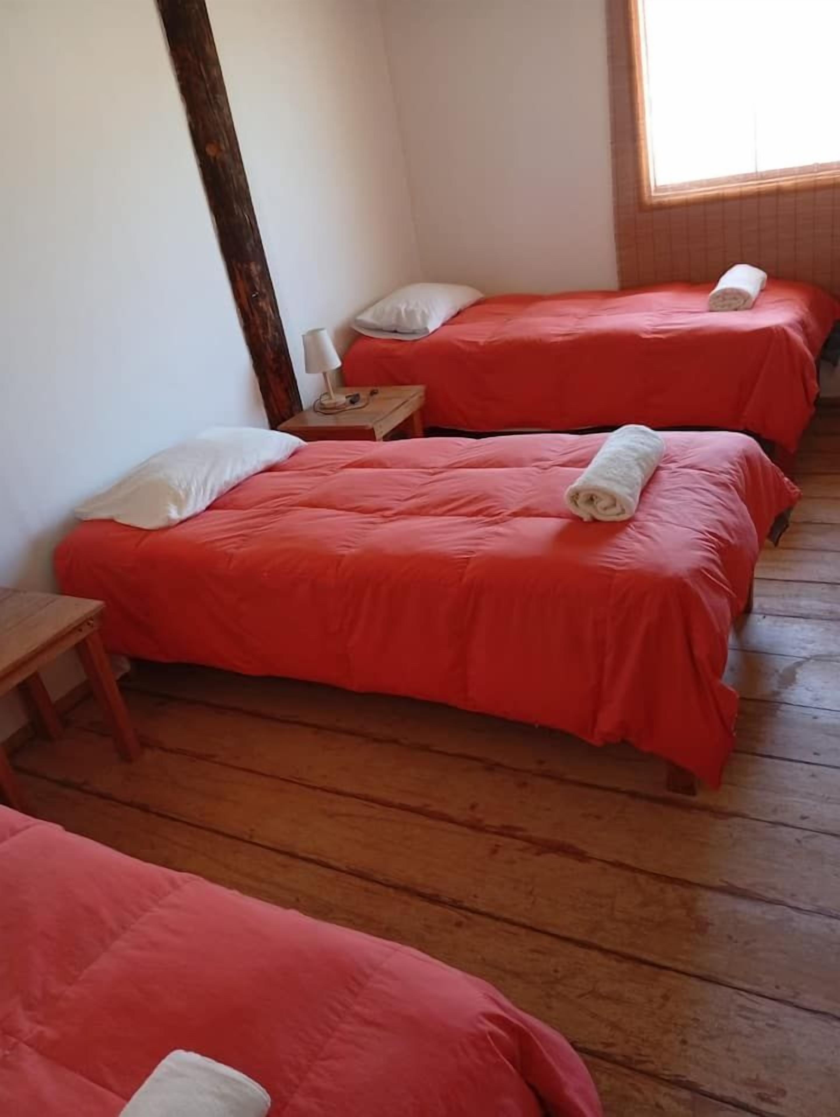 Una habitación con tres camas, cada una con una colcha roja y almohadas blancas. Hay un banco de madera y una mesita con una lámpara.