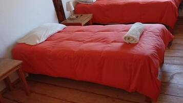 Una habitación con tres camas, cada una con una colcha roja y almohadas blancas. Hay un banco de madera y una mesita con una lámpara.