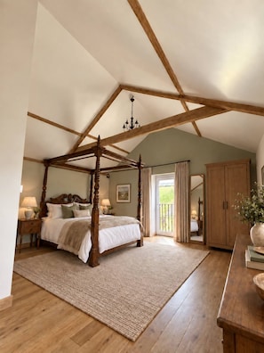 Un dormitorio con cama con dosel, vigas de madera, un candelabro, vistas a través de un ventanal y un armario de madera.