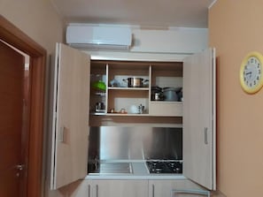 A compact kitchen with a gas stove, shelves with cookware, and a clock on the wall.