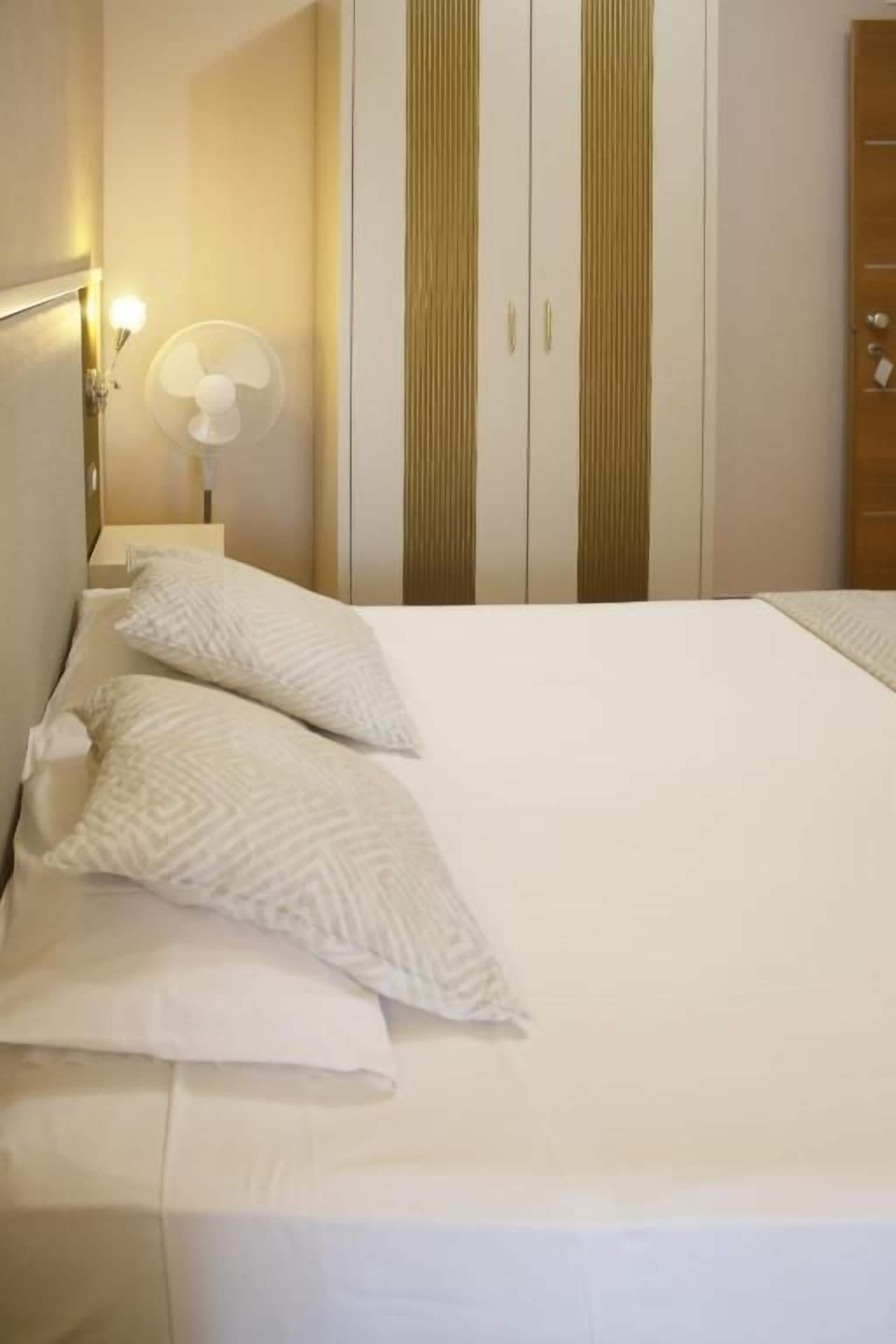 Una cama bien hecha con almohadas, una lámpara de noche y un ventilador de pared.