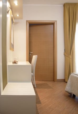 A room with a wooden door, a white chair, a small table with a glass container, and a bed with a white cover.