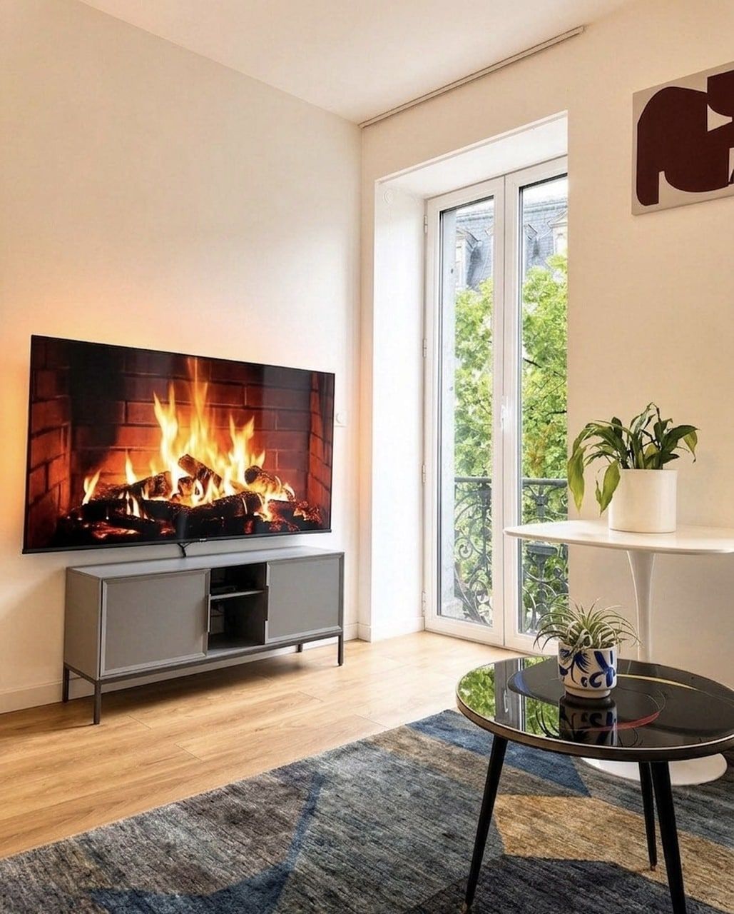 Una sala de estar con un televisor de pantalla plana montado en la pared, una chimenea con leña, un mueble de entretenimiento gris, una mesa de centro redonda de vidrio y una planta en maceta sobre un estante blanco.